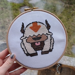 May include: A cross-stitch embroidery of Appa from Avatar: The Last Airbender. The design features Appa's face in brown, tan, and white, with an orange arrow on his head. The embroidery is set in a round wooden hoop.