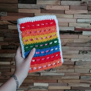 Op de afbeelding: Een regenboog gehaakte boekomslag met een witte rand. De omslag is gemaakt met verschillende gekleurde garens en heeft een gehaakt ontwerp.