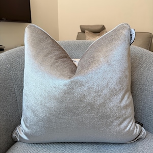 May include: A square, light gray velvet pillow with a soft sheen, resting on a gray upholstered chair. The pillow has a smooth texture and a subtle gradient of color, with a tag on the side.