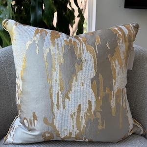 May include: A decorative square pillow with a textured design. The pillow features a gray background with gold and white abstract patterns. The pillow is placed on a gray armchair, with a plant and window in the background.