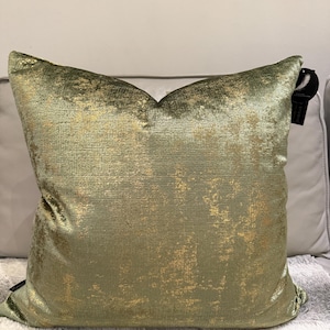 May include: A square decorative pillow with a textured, olive green fabric and gold accents. The pillow is on a gray surface and has a tag attached to the side.