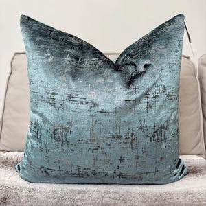 May include: A teal blue throw pillow with a metallic silver and black abstract pattern. The pillow is sitting on a light gray fuzzy surface.