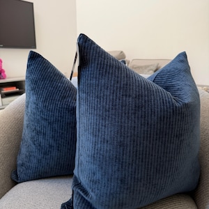 May include: Two navy blue corduroy throw pillows with vertical ribbed texture. The pillows are placed on a light gray upholstered chair, creating a contrast in textures and colors. The pillows have a soft, inviting appearance.