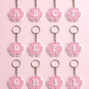 May include: A collection of twelve pink flower-shaped keychains, each with a different letter of the alphabet in white. The keychains have a silver chain and ring, and are set against a soft pink background.