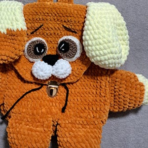 Könnte beinhalten: Ein brauner gehäkelter Rucksack in Form eines Hundes mit weißen Ohren und braunen Augen. Der Rucksack hat einen schwarzen Kordelzug und einen goldenen Metallverschluss.