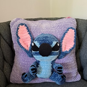 Op de afbeelding: Een handgemaakt pluche kussen met een blauw en lavendelkleurig Stitch-personage. Het kussen heeft grote, getextureerde oren met roze binnenkanten, zwarte ogen en een zwarte neus. Het kussen is ongeveer 40 cm vierkant.