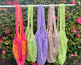 May include: A collection of five crocheted net bags in various colors, including a vibrant gradient of orange and pink, light green, purple, tan, and lime green. The bags are suspended from a silver bar against a backdrop of green foliage and pink flowers.