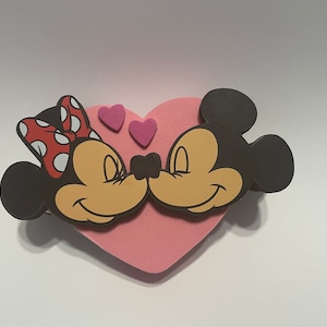 May include: A pink heart-shaped foam cutout with Mickey Mouse and Minnie Mouse kissing. Mickey is on the right and Minnie is on the left. They are both smiling with their eyes closed. There are three small pink hearts above them.