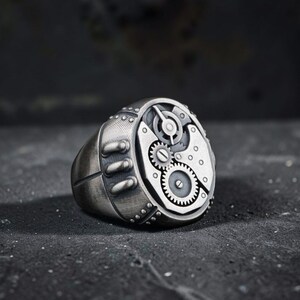 May include: A silver-colored ring with a steampunk design. The ring features a detailed mechanical design with visible gears, rivets, and a textured surface. The ring has a wide band and a circular face with intricate clockwork elements.