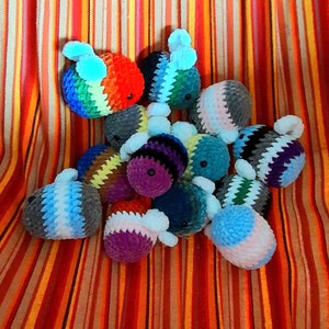 Può includere: Un gruppo di peluche di api all'uncinetto in vari colori, tra cui arcobaleno, viola, blu e grigio. I peluche sono disposti in una pila su uno sfondo a righe.