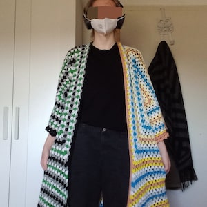 Könnte beinhalten: Ein langer, gehäkelter Cardigan mit einem schwarz-weißen Streifenmuster auf der einen Seite und einem mehrfarbigen Streifenmuster auf der anderen Seite. Der Cardigan wird über einem schwarzen T-Shirt und einer schwarzen Hose getragen.