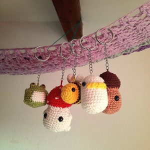 Könnte beinhalten: Fünf gehäkelte Tier-Schlüsselanhänger, die an silbernen Schlüsselringen hängen. Die Schlüsselanhänger sind ein grüner Frosch, ein roter und weißer Pilz, eine gelbe und schwarze Biene, ein weißer Vogel und eine braune Eichel.