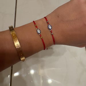 May include: A gold-colored bracelet and two red string bracelets with blue and white evil eye charms. The gold bracelet is a cuff style. The evil eye bracelets have gold-colored beads.