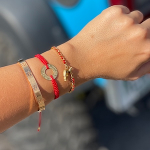 May include: A woman's wrist with three bracelets. The bracelets are a gold chain bracelet, a red string bracelet with a gold circle charm, and a gold bangle bracelet with the inscription "LOVE".