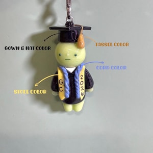 May include: A pale green figurine wearing a black graduation cap with a gold tassel and a black gown. A yellow stole features the letters GC on one side and the year 2026 on the other, accented by a blue braided cord. The item includes a metal keychain.