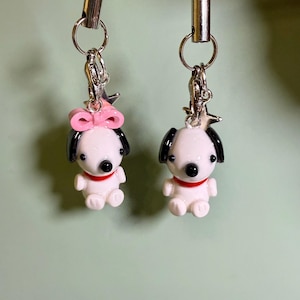 May include: Two white dog-shaped charms with black ears and eyes, and a red collar. One charm has a pink bow. Each charm is attached to a silver-colored clasp and black cord. The charms are small, likely under 2 inches in height.
