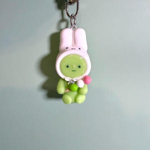 May include: A green figurine keychain of a character wearing a white bunny hood. The character holds a pink and green lollipop. The keychain has a silver clasp and chain.