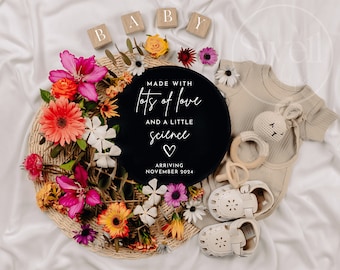 May include: A black and white round sign with the text "Made with lots of love and a little science" and a heart. The sign is surrounded by colorful flowers and a baby outfit with a bunny rattle.