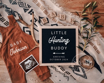 May include: A brown baby onesie with the text "Baby Johnson" on the pocket, a black and white ultrasound image, a wooden sign with the text "Little Hunting Buddy Arriving October 2024", and wooden blocks spelling "BABY" on a white and black patterned blanket.