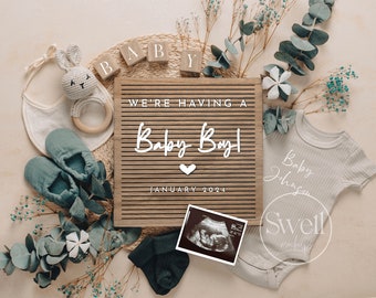 以下が含まれることがあります： 「We're having a Baby Boy! January 2024」と書かれた木製のレターボード。周りには、ガラガラ、ベビーシューズ、ロンパース、ユーカリの葉など、赤ちゃん用品が置かれています。
