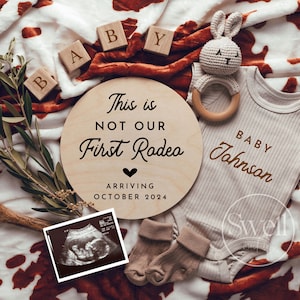 May include: A wooden round sign with the text "This is not our first rodeo" and a heart symbol. The sign also includes the text "Arriving October 2024". A baby onesie with the text "Baby Johnson" is visible in the image. The image also includes a stuffed bunny, a wooden baby rattle, and a sonogram image.