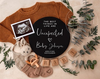 May include: A black chalkboard sign with the text "The best things in life are unexpected" and a heart symbol. Below the text is "Baby Johnson" and "Arriving November". The sign is surrounded by a brown knitted sweater, a small knitted bunny toy, a wooden baby rattle, and wooden blocks that spell out "BABY".