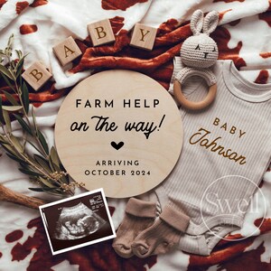 Könnte beinhalten: Ein Holzschild mit dem Text "FARM HELP on the way!" und "ARRIVING OCTOBER 2024". Ein Baby-Strampler mit dem Text "BABY Johnson" liegt auf einer braunen und weißen Kuhfelldecke. Es gibt Holzklötze, die "BABY" buchstabieren, und ein gehäkeltes Hasenrassel.