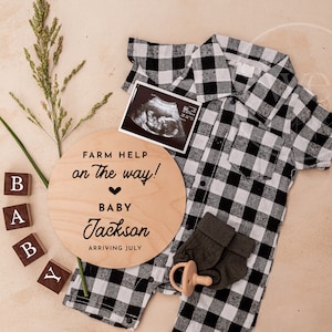 Könnte beinhalten: Ein Holzschild mit dem Text "Farm Help on the way!" und einem Herz mit dem Text "Baby Jackson Arriving July". Das Schild ist umgeben von einem schwarz-weiß karierten Baby-Outfit, einem Schnuller und Holzklötzen, die "BABY" buchstabieren.
