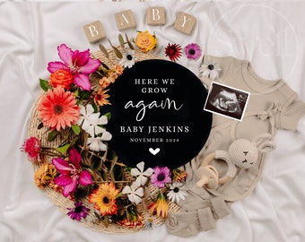 May include: A black and white round sign with the text "Here we grow again" and "Baby Jenkins November 2024". The sign is surrounded by a wreath of colorful flowers and a baby onesie with an ultrasound image.