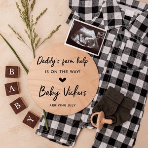 Könnte beinhalten: Ein rundes Schild aus Holz mit dem Text "Daddy's farm help is on the way! Baby Vickers arriving July" mit einem Herzsymbol. Das Schild ist umgeben von Holzblöcken, die "BABY" buchstabieren, und einem schwarz-weiß karierten Hemd mit einem Paar grüner Socken und einem Schnuller.