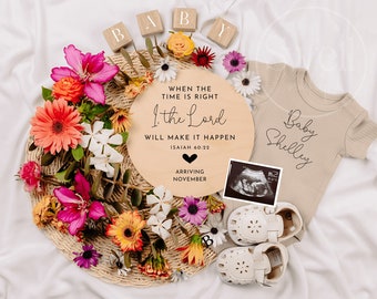 May include: A wooden round sign with the text "When the time is right, I, the Lord will make it happen, Isaiah 60:22" and a black heart. The sign also says "Arriving November". A baby onesie with the text "Baby Shelley" is in the background. The sign is surrounded by flowers and a baby's shoes.