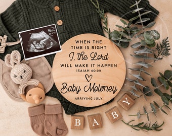 May include: A wooden round sign with the text "WHEN THE TIME IS RIGHT I. the Lord WILL MAKE IT HAPPEN ISAIAH 60:22" and a heart symbol. Below the text is "Baby Moloney ARRIVING JULY".  The sign is surrounded by greenery and a few wooden blocks that spell out "BABY".