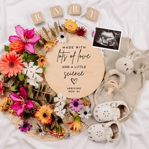 May include: A wooden round sign with the text "Made with lots of love and a little science" with a heart symbol and the date "Nov '24". The sign is surrounded by flowers, a baby outfit, a rattle, and a pair of baby shoes.