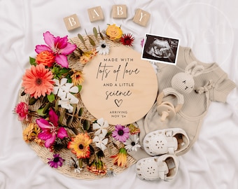 May include: A wooden round sign with the text "Made with lots of love and a little science" with a heart symbol and the date "Nov '24". The sign is surrounded by flowers, a baby outfit, a rattle, and a pair of baby shoes.