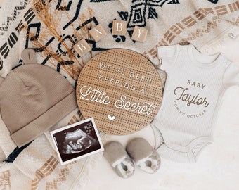 Könnte beinhalten: Ein Schild aus Holz mit dem Text "We've Been Keeping a Little Secret" und einem weißen Herz. Ein weißer Strampler mit dem Text "Baby Taylor Coming October" in brauner Schrift. Eine braune Strickmütze, ein Paar braune Booties und ein Schwarzweiß-Ultraschallbild sind ebenfalls im Bild enthalten.