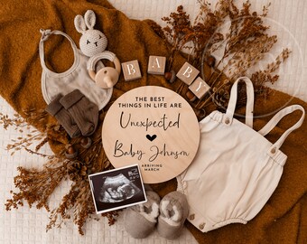 May include: A wooden round sign with the text "The best things in life are unexpected" and "Baby Johnson arriving March". The sign is surrounded by a brown blanket, a baby onesie, a pair of booties, a stuffed bunny, a wooden block spelling "BABY", and a sonogram image.