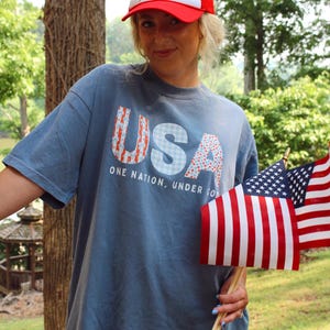 Könnte beinhalten: Ein blaues T-Shirt mit dem Wort "USA" in rot-weiß-blauen Mustern. Der Text "ONE NATION, UNDER GOD" steht darunter. Ein rot-weißer "God Bless America" Hut wird getragen. Amerikanische Flaggen werden gehalten.