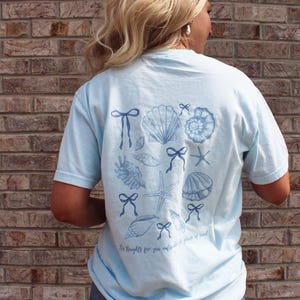May include: Light blue t-shirt featuring a collection of seashells, bows, and starfish in a darker blue hue. The shirt has the text "My thoughts for you outside the power of reach" printed at the bottom. The shirt is a casual wear item.