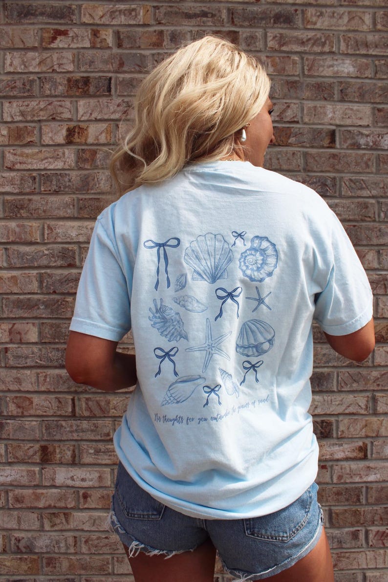 May include: Light blue t-shirt featuring a collection of seashells, bows, and starfish in a darker blue hue. The shirt has the text "My thoughts for you outside the power of reach" printed at the bottom. The shirt is a casual wear item.