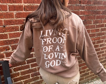 Op de afbeelding: Een bruine sweatshirt met de tekst "LIVING PROOF OF A LOVIN GOD" in witte letters. De persoon draagt een beige broek. De achtergrond is een rode bakstenen muur.