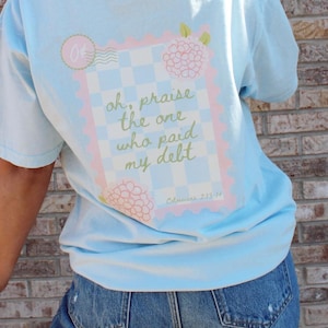 Könnte beinhalten: Hellblaues T-Shirt mit einem Aufdruck auf der Rückseite, der ein Briefmarkendesign zeigt. Die Briefmarke enthält den Text "oh, praise the one who paid my debt" und eine Blumenillustration. Das Shirt wird mit blauen Jeans getragen.