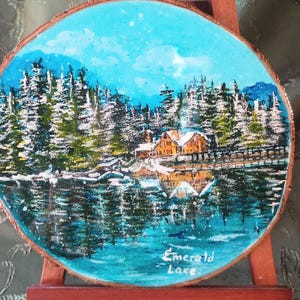 May include: Oval painting of a winter scene with a lake and snow-covered trees, mounted on a small wooden easel. The artwork is titled "Emerald Lake".