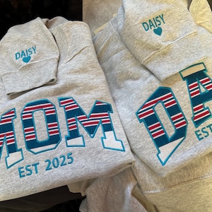 May include: Gray sweatshirts with embroidered text. One says "MOM EST 2025" in teal, red, and white. Another says "DAD EST 2025" in the same colors. The sleeve of one sweatshirt has "DAISY" and a teal heart.