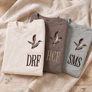 May include: Three crewneck t-shirts in cream, brown, and sage green, each featuring a chest pocket embroidered with a flying mallard duck and a three-letter monogram. The cream shirt has dark brown letters, the brown shirt has tan letters, and the sage shirt has dark brown letters.