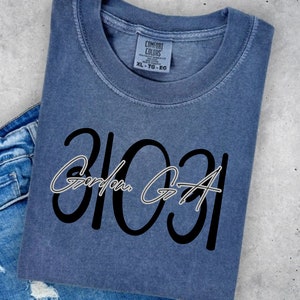 May include: A blue Comfort Colors t-shirt with the text "31031 Gordon GA" in black and white. The shirt is folded and placed on a light gray surface with a pair of blue jeans.