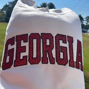 Op de afbeelding: Een witte sweatshirt met het woord "GEORGIA" in grote, rode, glitterende letters met een zwarte omlijning. Het kledingstuk wordt gepresenteerd tegen een achtergrond van blauwe lucht en groen gras.