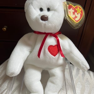 Puede incluir: Oso de peluche blanco Beanie Baby con una cinta roja y un corazón rojo en el pecho. El oso tiene ojos marrones y una nariz marrón. El oso está sentado en una vitrina de plástico transparente. La etiqueta dice "Beanie Baby".
