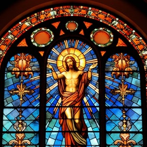 May include: A stained glass window depicting Jesus Christ with outstretched arms, surrounded by a halo and rays of light. The window is set in a church or cathedral, with a blue and gold background.