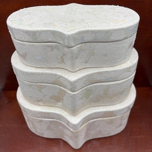 May include: A stack of three white, heart-shaped ceramic boxes with iridescent accents. The boxes have a textured, speckled surface and a glossy finish. The lids fit snugly, creating a cohesive set for storage or display.
