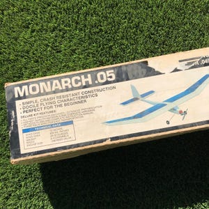 May include: A box for a Monarch .05 model airplane kit. The box is brown with blue and white text. The text on the box reads "Monarch .05", "Simple, Crash Resistant Construction", "Docile Flying Characteristics", "Perfect for the Beginner", "Deluxe Kit Features", and "Technical Data". The box also features a picture of the airplane.